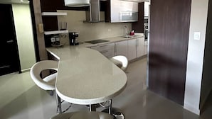 Uma cozinha moderna com ilha central, cadeiras brancas, eletrodomésticos de aço inoxidável e uma porta de madeira escura.