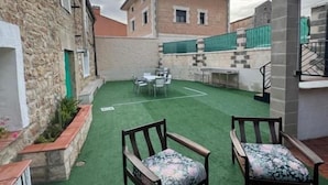 A courtyard with artificial turf, two wooden chairs, a table, and a building in the background.
