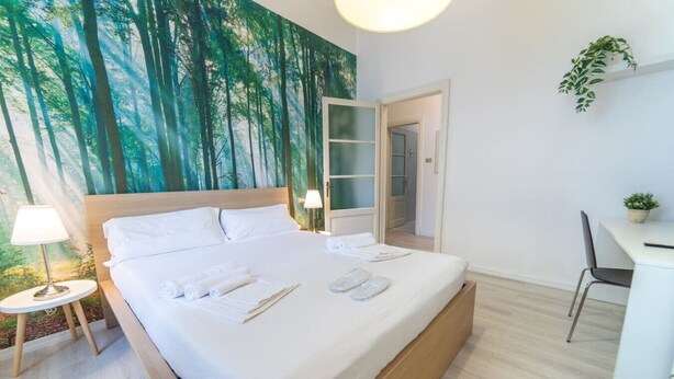 A neatly made bed with white linens and towels, a small bedside table with a lamp, a desk with a chair, and a green plant on the wall. - Cool stay at 2 steps from Duomo, Como (Como)