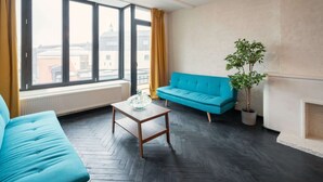 A room with a large window, a blue sofa, a wooden coffee table, and a potted plant. - Comfortable 2br/14p Apartment - Central Antwerp (Antwerp)