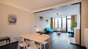 A room with a wooden dining table, white chairs, a blue sofa, and a window with curtains. - Comfortable 2br/14p Apartment - Central Antwerp (Antwerp)