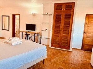 Un dormitorio con cama, escritorio, televisor, armario y un baño visible a través de una puerta abierta.