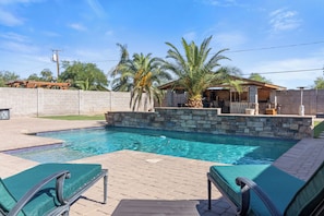 A swimming pool with a stone border, two lounge chairs, and a palm tree.