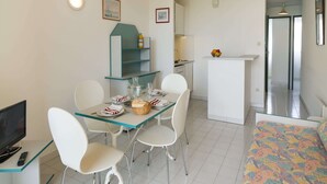 Un comedor con una mesa de vidrio, sillas blancas, un televisor y una cocina al fondo.