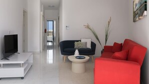Una sala moderna con un televisor de pantalla plana sobre un soporte blanco, un sofá azul oscuro con un cojín blanco, un sofá rojo y una mesa redonda de centro con un jarrón y una planta.