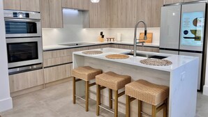 Cozinha moderna com armários de madeira, eletrodomésticos de aço inoxidável, uma ilha branca com bancos e iluminação pendente.