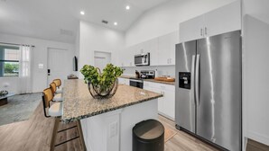 A modern kitchen with white cabinets, a stainless steel refrigerator, a granite countertop, and a small island with chairs. - Emerald Entry Retreat (LaBelle)
