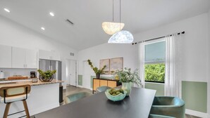 A modern kitchen with white cabinets, a central island, and a dining area with a dark table and teal chairs. - Emerald Entry Retreat (LaBelle)