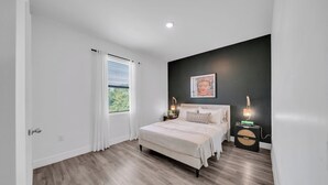A bedroom with a bed, a nightstand, a wall-mounted lamp, and a window with curtains. - Emerald Entry Retreat (LaBelle)