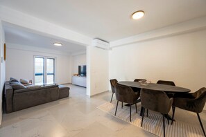 Een moderne woonkamer met een bank, eettafel en stoelen, een keuken met een televisie en een groot raam met zonwering.