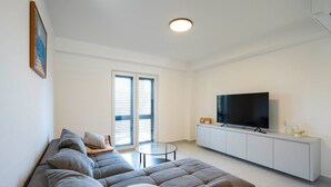 A modern living room with a grey sectional sofa, a glass coffee table, a flat-screen TV on a white cabinet, and a large window with blinds. - #1 Holiday Apartment MTG - Orebić (Orebić)