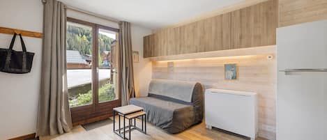 A compact room with a sofa, a small table, a refrigerator, and a window with curtains.