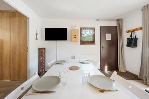A dining area with a long table set for four, a television mounted on the wall, a window with a view of a forest, and a door with a coat rack. - 'Parenthèse Alpine' Apartment with Mountain View and Private Terrace (La Plagne-Tarentaise)