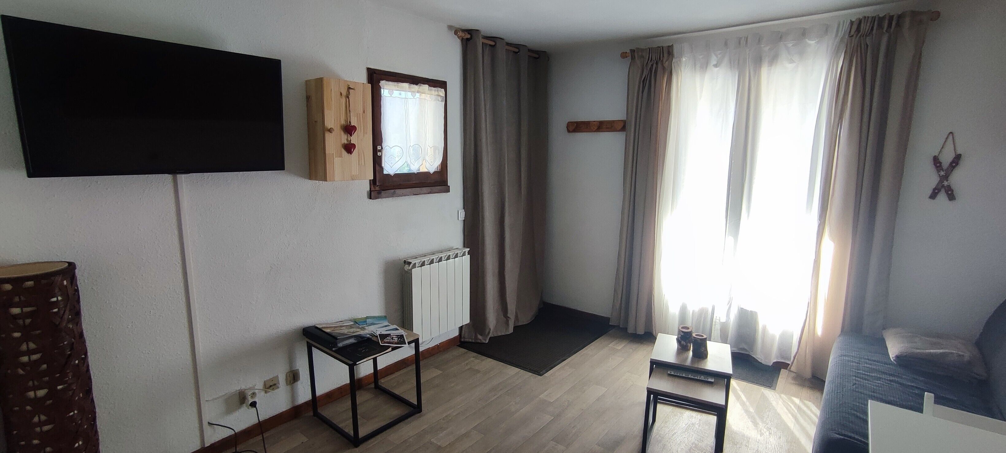 Una sala de estar con una televisión de pantalla plana, un mueble de madera con un ventanal, un radiador, una mesita con revistas, un sofá y un ventanal con cortinas.