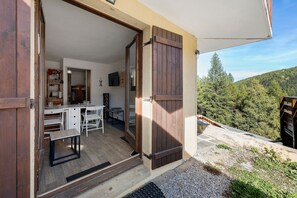 An open door leading to a dining area with a table and chairs, a kitchen area with cabinets and a sink, and a view of a forested hillside. - 'Parenthèse Alpine' Apartment with Mountain View and Private Terrace (La Plagne-Tarentaise)