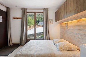 A bedroom with a bed, curtains, a wooden headboard, and a view of a wooden deck and trees. - 'Parenthèse Alpine' Apartment with Mountain View and Private Terrace (La Plagne-Tarentaise)