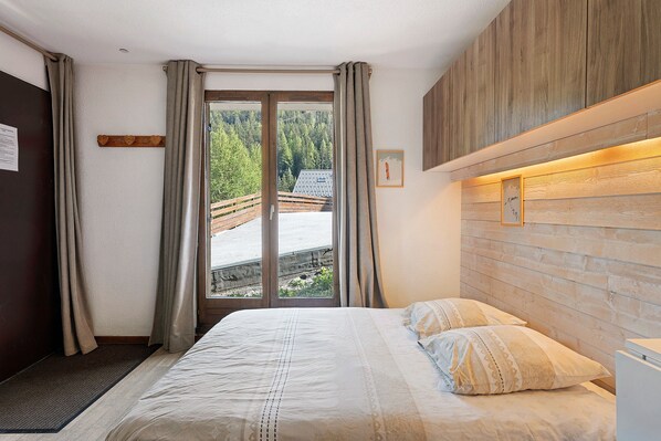 Un dormitorio con cama, cortinas, cabecera de madera y vistas a una terraza de madera y árboles.