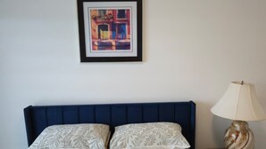 Ein Bett mit blauem Kopfteil, einer gemusterten Bettdecke, zwei Kissen, einem gerahmten Kunstwerk darüber und einer Nachttischlampe auf einem Holznachttisch.