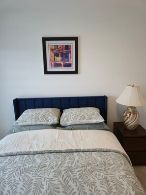 Een bed met een blauw hoofdbord, een bed met een decoratief patroon, twee kussens, een ingelijst kunstwerk erboven en een bedlamp op een houten nachtkastje.