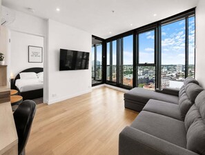 A modern living room with a large window, a flat-screen TV, a sofa, and a bed. - 2BR Stay in Fortitude Valley w/ pool and gym (Brisbane)