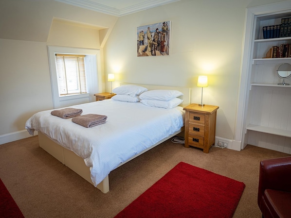 A bedroom with a bed, two bedside tables, a red rug, a window with blinds, a painting on the wall, and a bookshelf.
