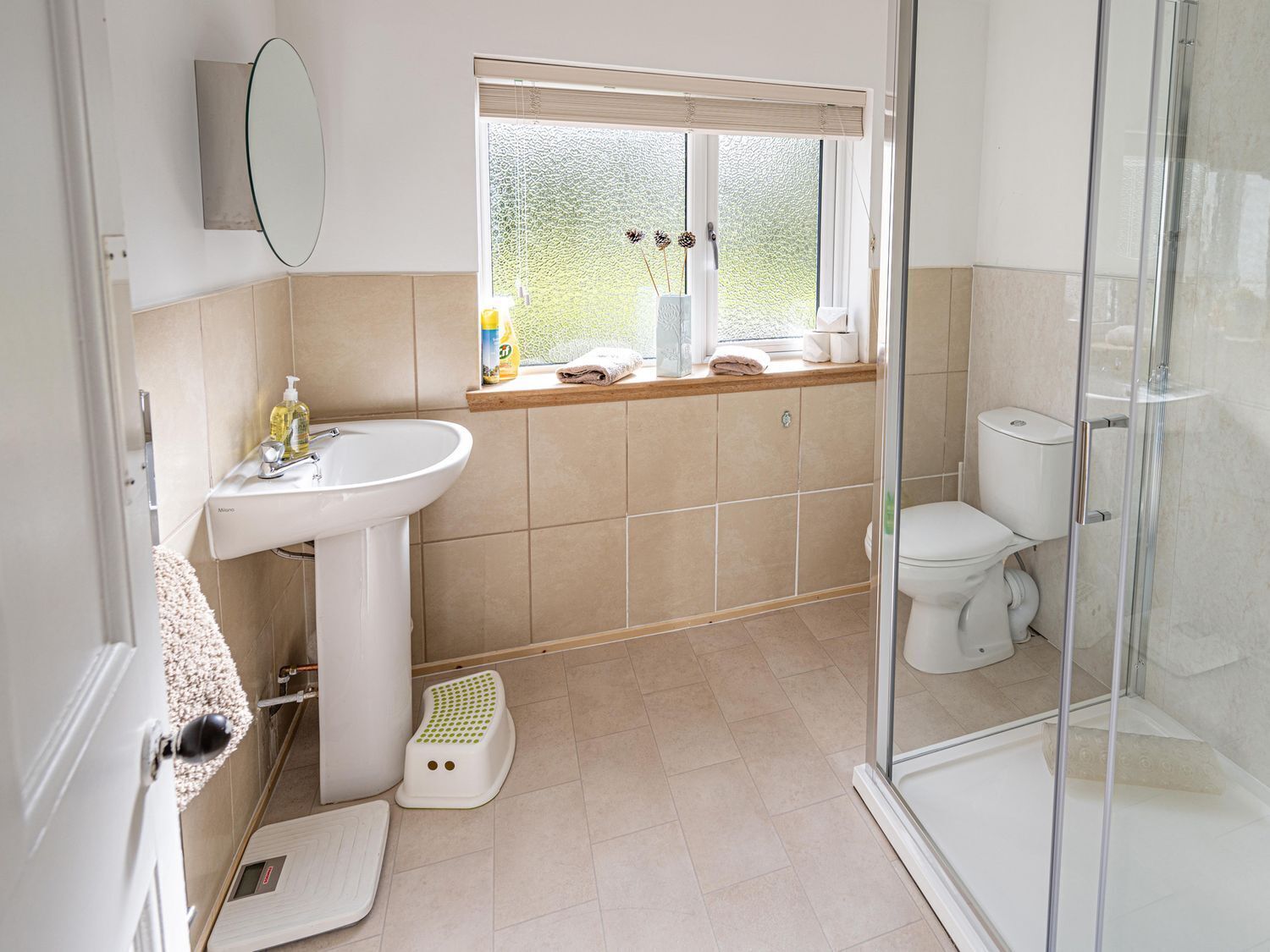 A bathroom with a sink, toilet, shower, and a window with a view of greenery.