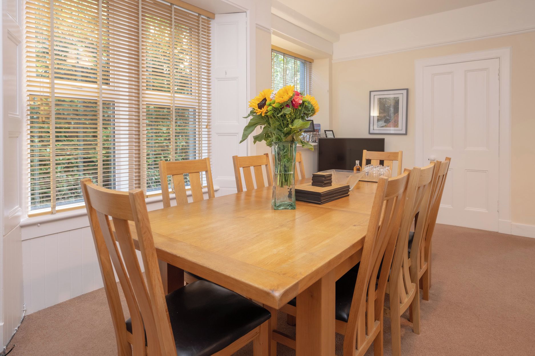Un comedor con una mesa de madera, sillas, un florero con flores y un televisor sobre un mueble bajo.