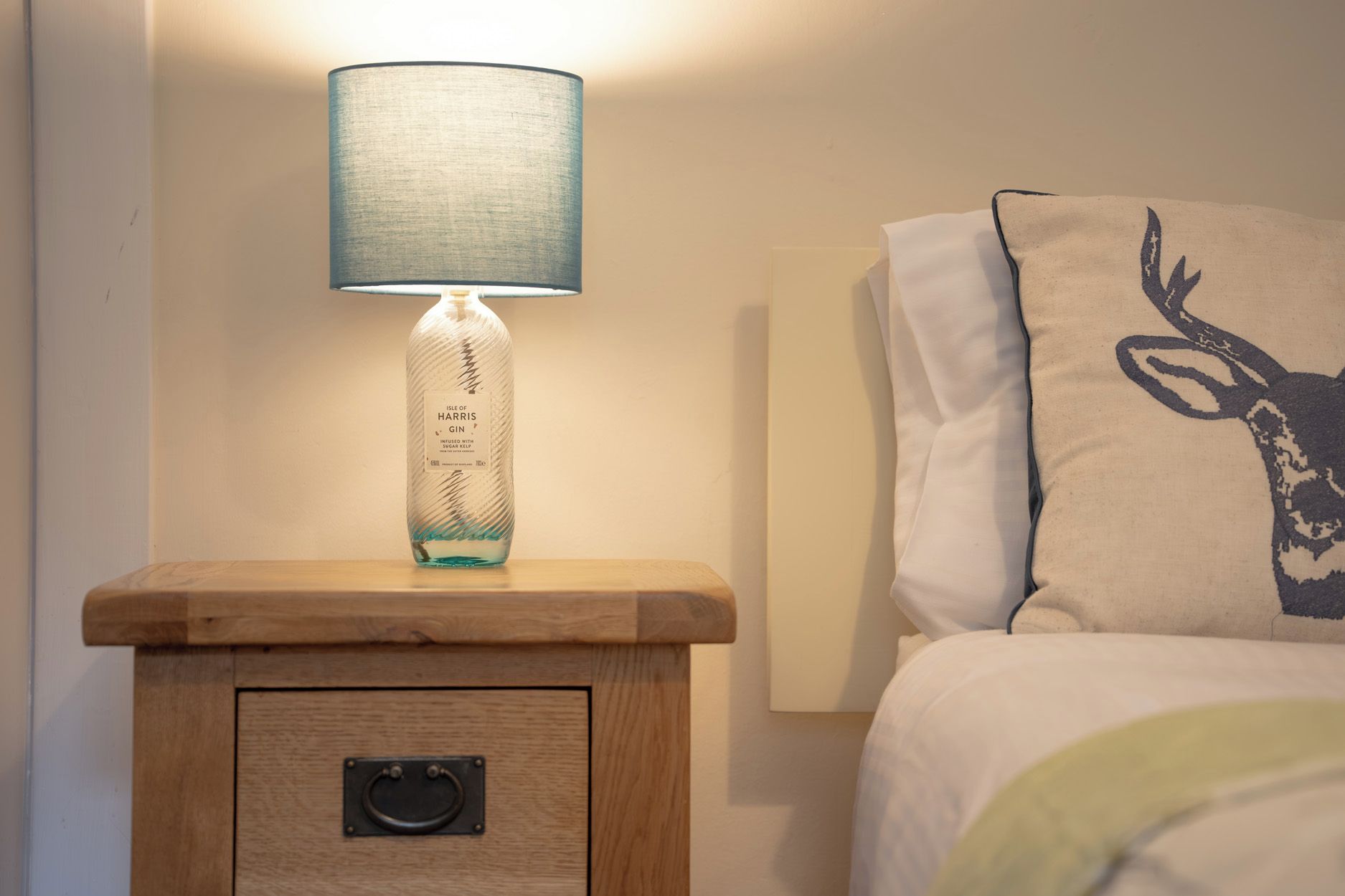 Une table de chevet avec une lampe et une bouteille de gin, un lit avec un oreiller orné d’un motif de cerf, et une petite table en bois avec un tiroir.