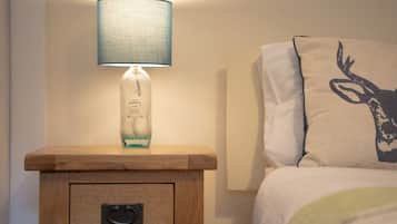 Une table de chevet avec une lampe et une bouteille de gin, un lit avec un oreiller orné d’un motif de cerf, et une petite table en bois avec un tiroir.