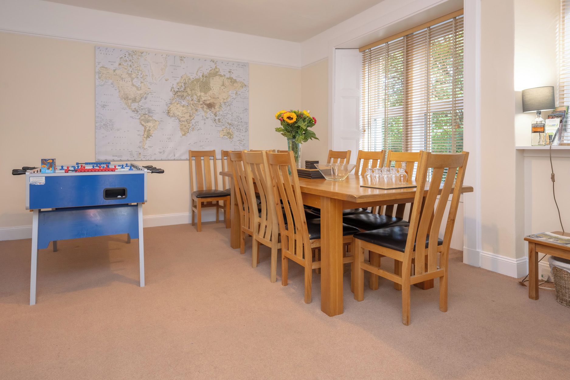 Un área de comedor con una mesa y sillas de madera, una mesa de fútbol de mesa, un mapa mundial en la pared y una ventana con persianas.