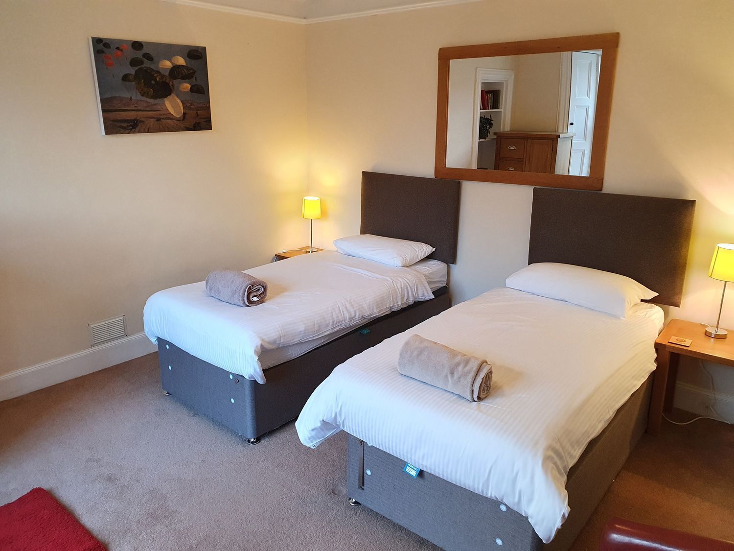 A hotel room with two beds, a mirror, a painting, and a red rug.