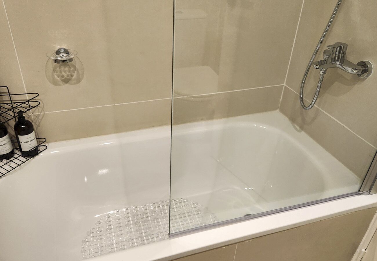 Casa de banho moderna com banheira de hidromassagem separada por vidro, chuveiro e suporte para garrafas com artigos de higiene pessoal.