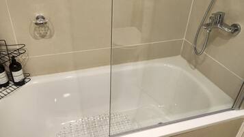 Casa de banho moderna com banheira de hidromassagem separada por vidro, chuveiro e suporte para garrafas com artigos de higiene pessoal.