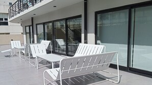 Outdoor seating with metal frames and white cushions, a glass door entrance, and a building with a balcony.