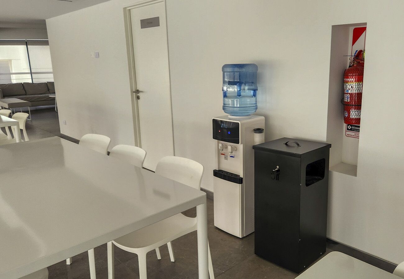 A conference room with a long table, white chairs, a water dispenser, a fire extinguisher, and a small trash bin.