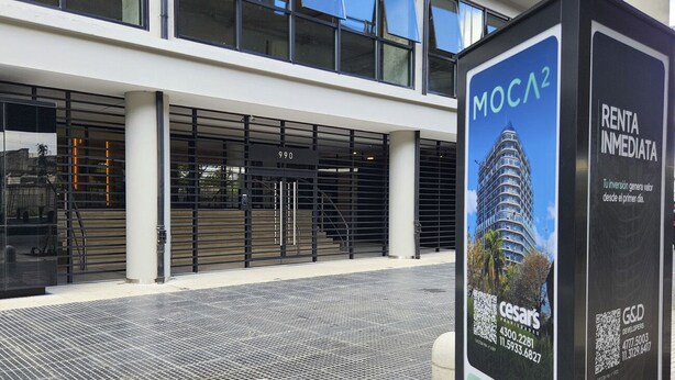 A modern building with a glass facade, a metal entrance gate, and a signboard advertising a property named 'MOCA2' with contact information.