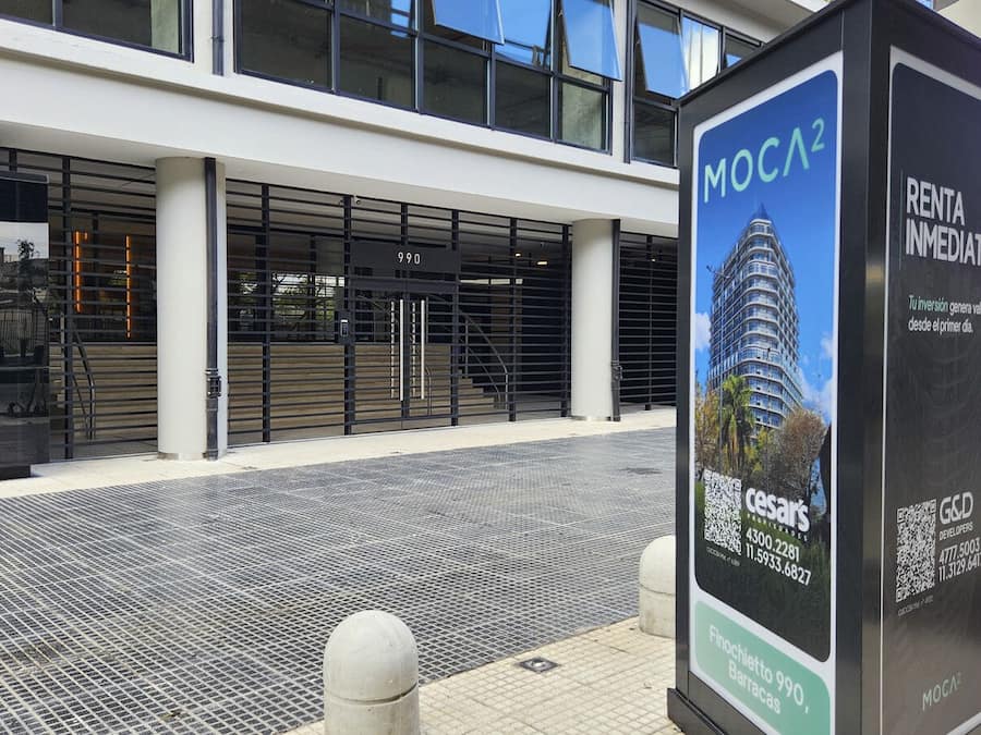 Um edifício moderno com fachada de vidro, portão de entrada metálico e uma placa indicando uma propriedade chamada 'MOCA2' com informações de contato.