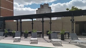 A poolside area with lounge chairs and tables, a clear pool, and a building in the background.