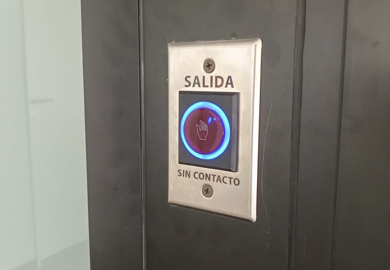 Un botón de ascensor metálico con los textos 'SALIDA' y 'SIN CONTACTO', e icono de mano que indica que no es necesario contacto.