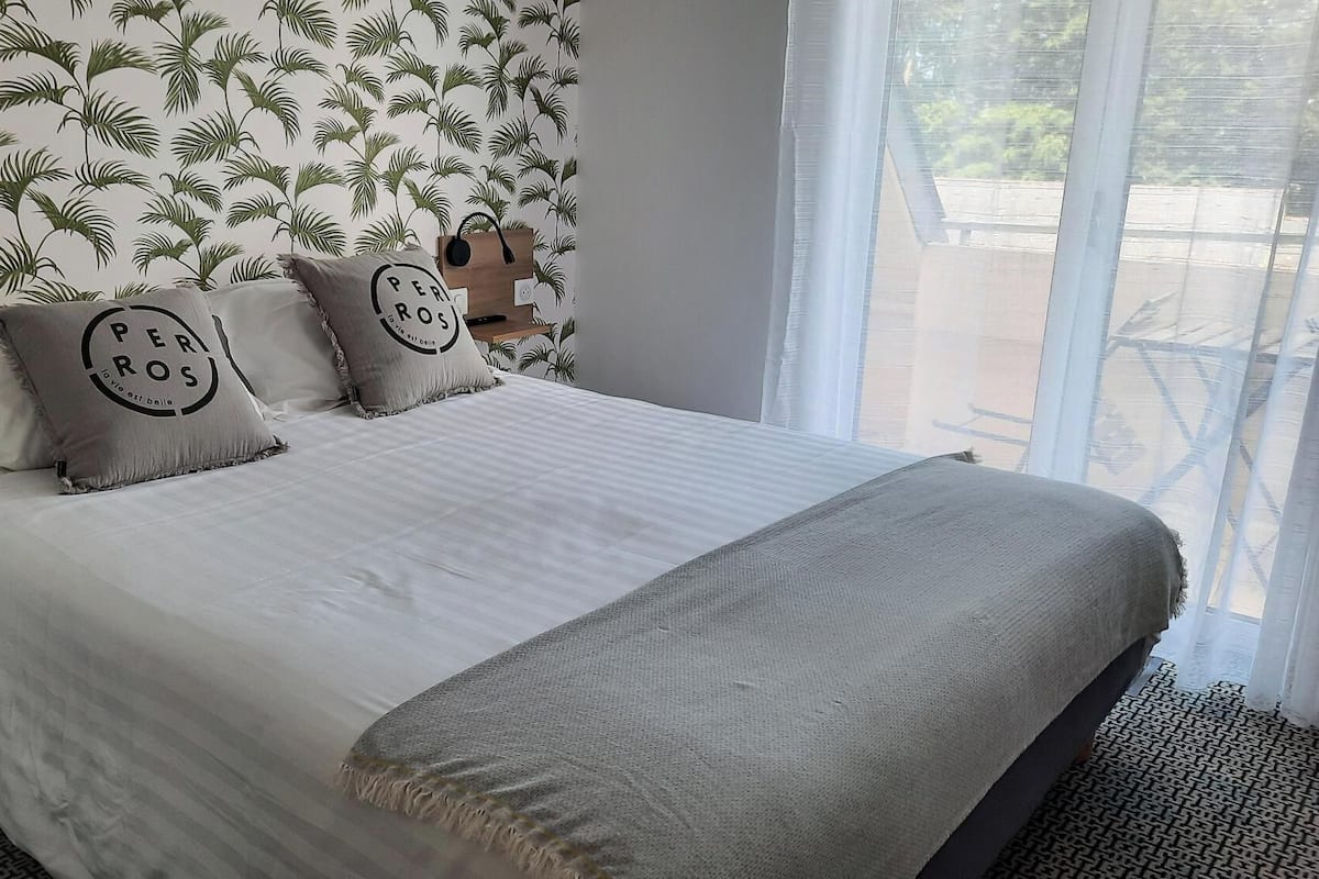 A bed with a white bedspread, two pillows with 'PER ROS' text, and a grey throw blanket.