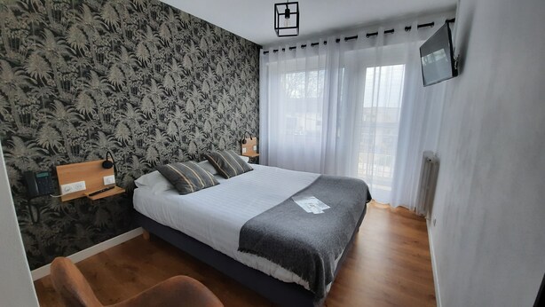 Un dormitorio con cama, escritorio con teléfono, televisión en la pared y ventana con cortinas.