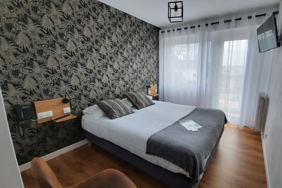 A bedroom with a bed, a desk with a phone, a TV on the wall, and a window with curtains.