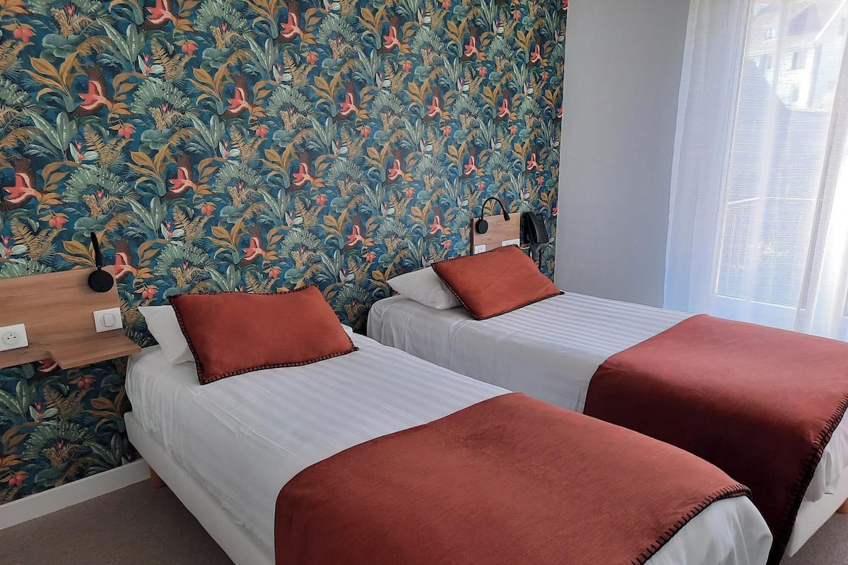A hotel room with two beds, a wooden headboard, a wall with a floral pattern, and a window with curtains.