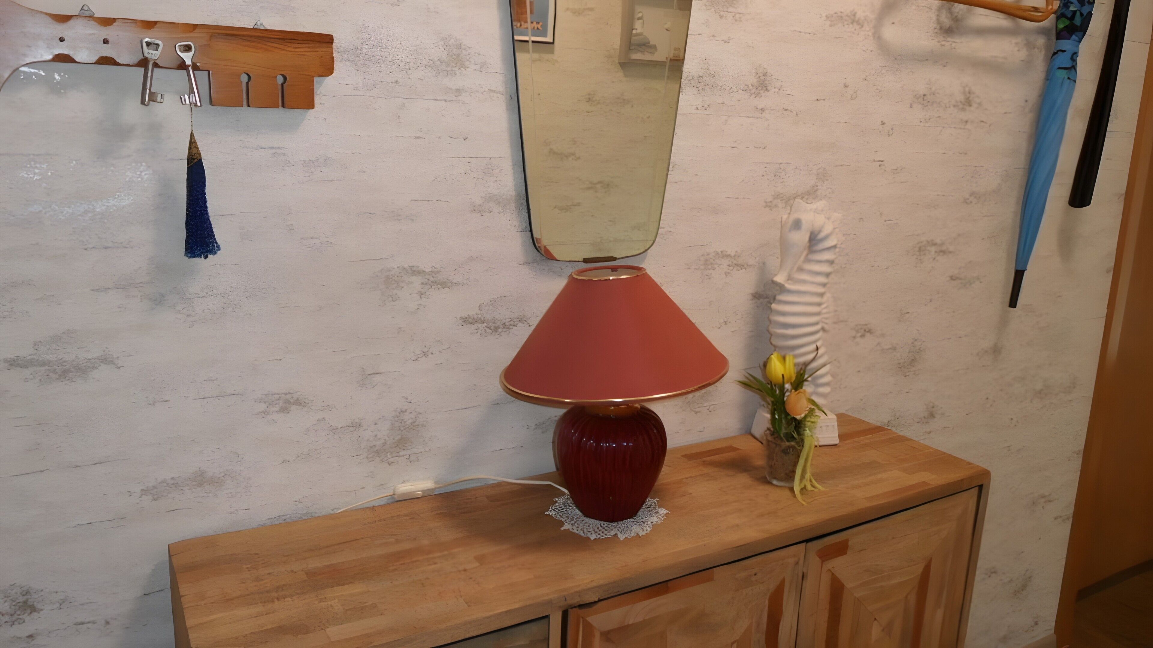 Um console de madeira com uma luminária vermelha, um vaso com flores amarelas e um espelho em cima.