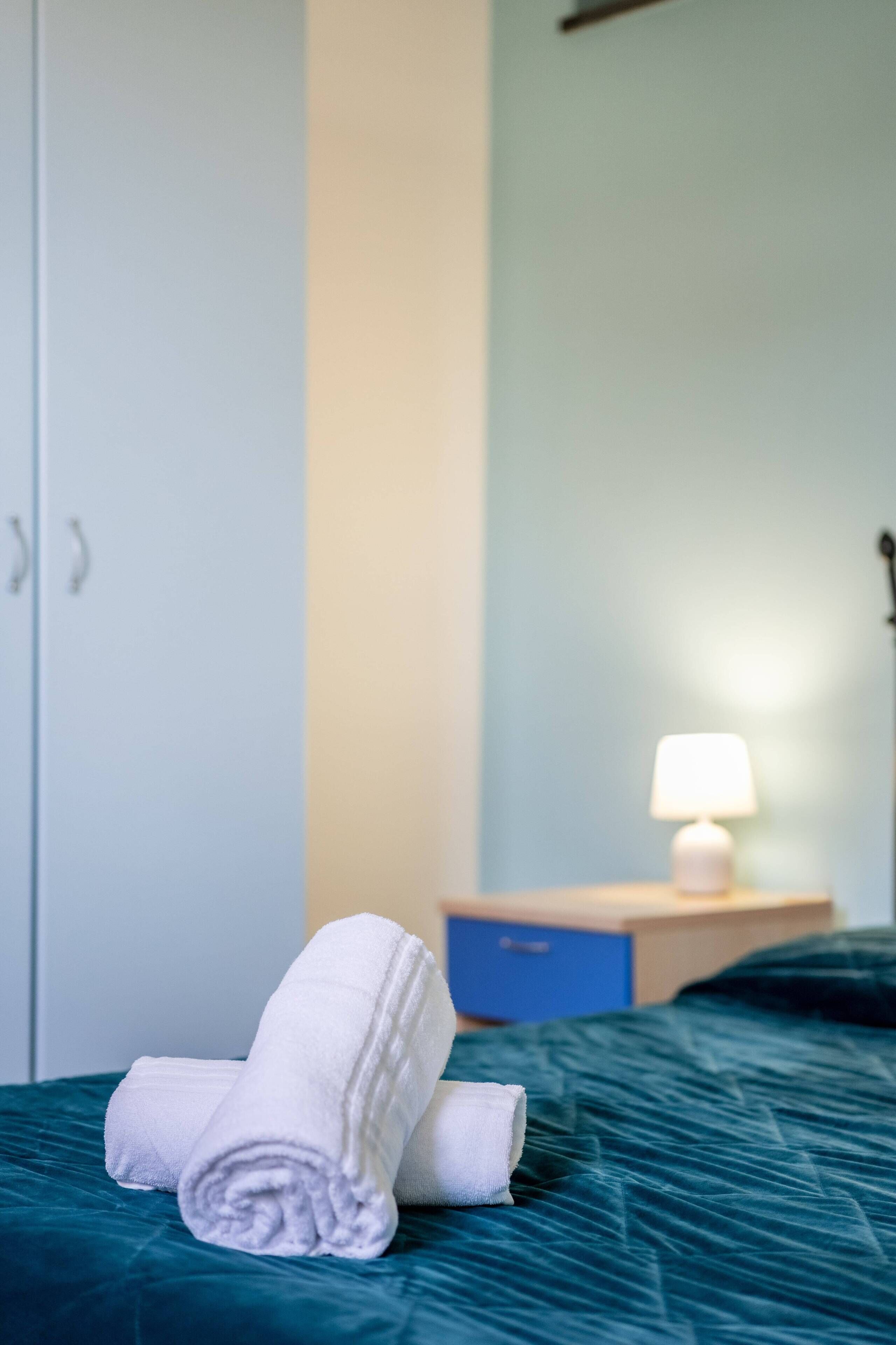 Una cama bien hecha con toallas blancas enrolladas, una mesita de noche azul con una lámpara y un armario.