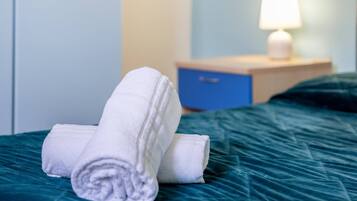 Una cama bien hecha con toallas blancas enrolladas, una mesita de noche azul con una lámpara y un armario.