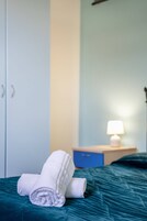 Una cama bien hecha con toallas blancas enrolladas, una mesita de noche azul con una lámpara y un armario.
