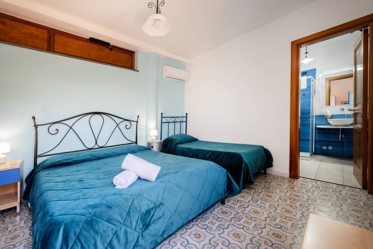 Un dormitorio con dos camas, una mesita de noche, una lámpara y un baño visible a través de una puerta abierta.