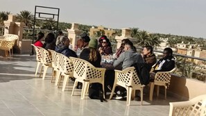 A group of people seated on plastic chairs outdoors with a tiled floor, palm trees, and buildings in the background.
