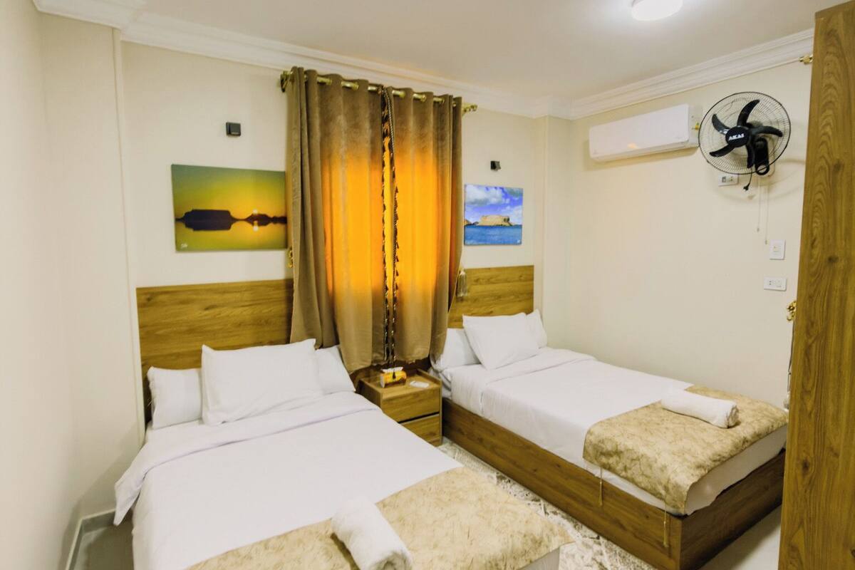 A hotel room with two beds, a nightstand, a wall-mounted fan, and a painting of a sunset.